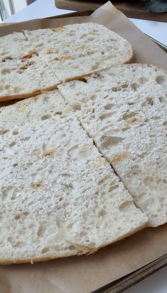 Four pieces of rustic, lightly floured bread halves are laid out on parchment paper, showing a soft, airy, and open crumb structure.
