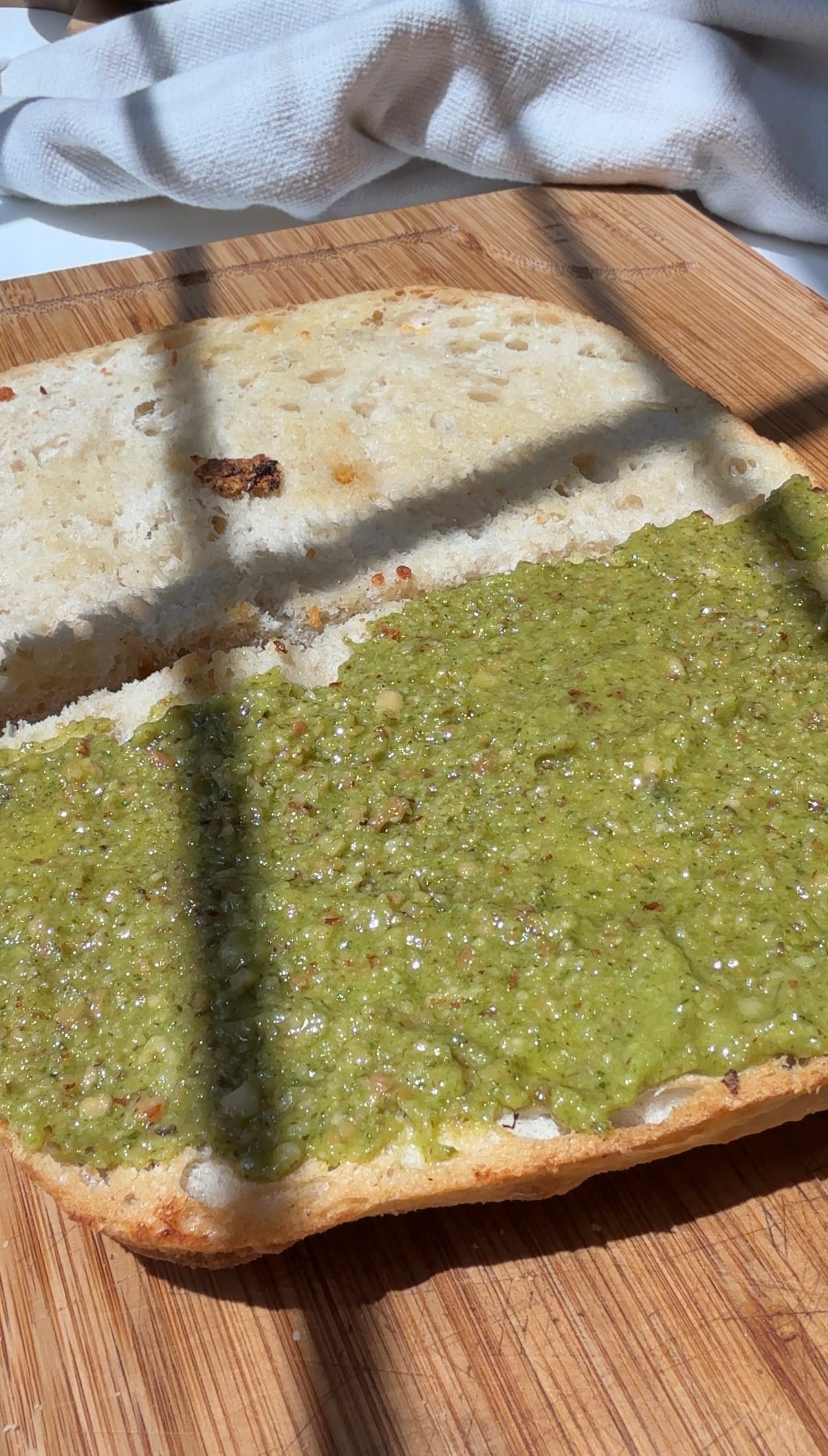 A sandwich bun cut in half on a wooden board, with green pesto spread on the bottom half. Sunlight and shadows cross the bread.