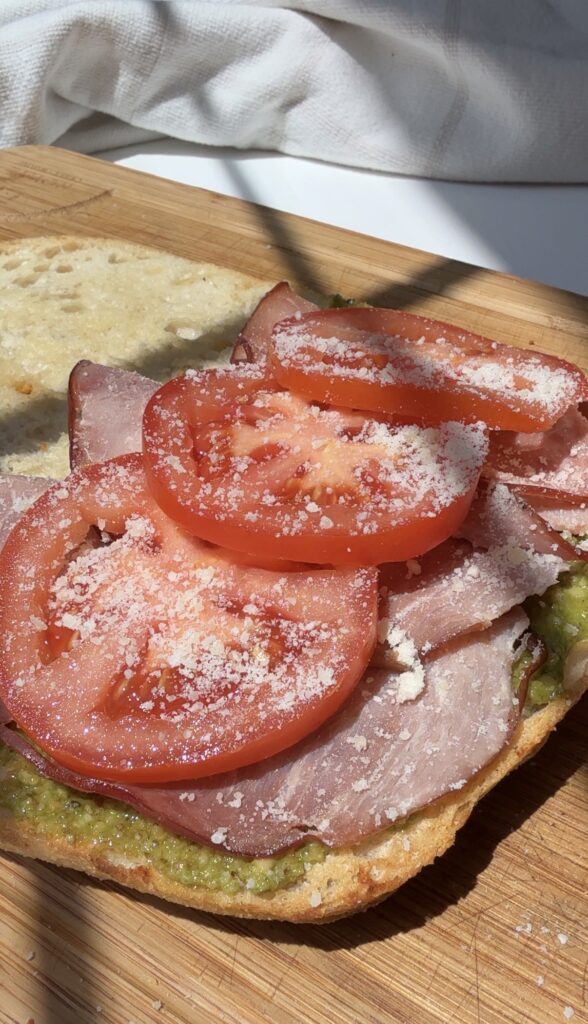 An open-faced sandwich with sliced ham, tomato, grated cheese, and green spread on bread sits on a wooden cutting board in sunlight.