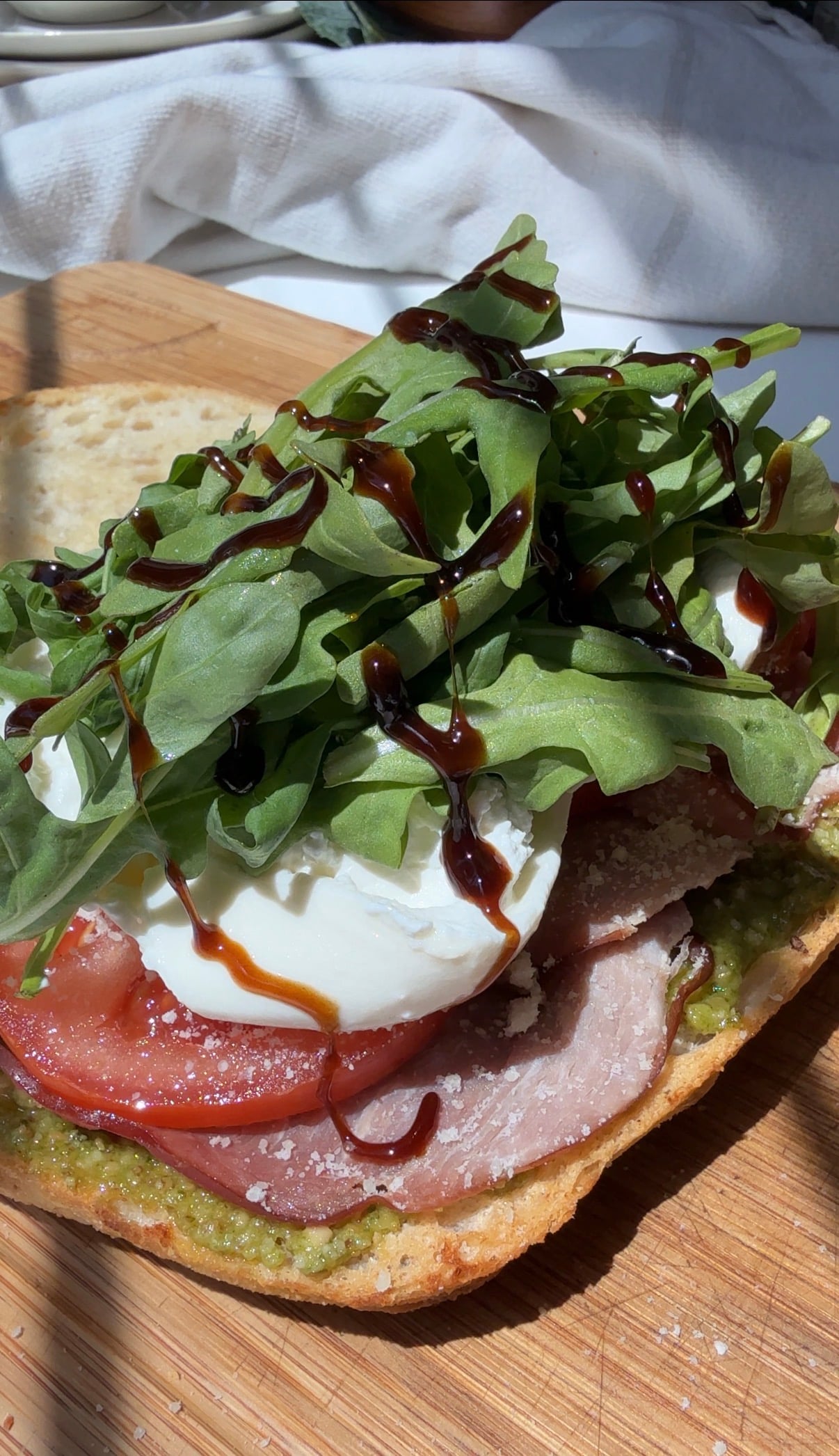 Open-faced sandwich with ham, tomato, fresh mozzarella, arugula, pesto, and a drizzle of balsamic glaze on toasted bread, on a wooden board.