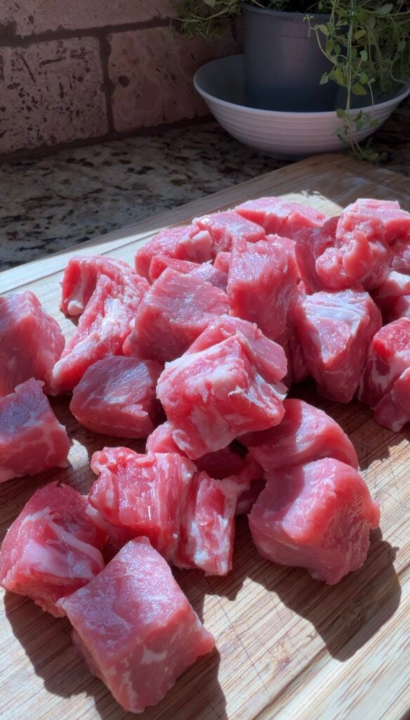 Chunks of raw, red meat with visible marbling are arranged on a wooden cutting board, brightly lit by sunlight from a nearby window. In the background, kitchen items are slightly out of focus.