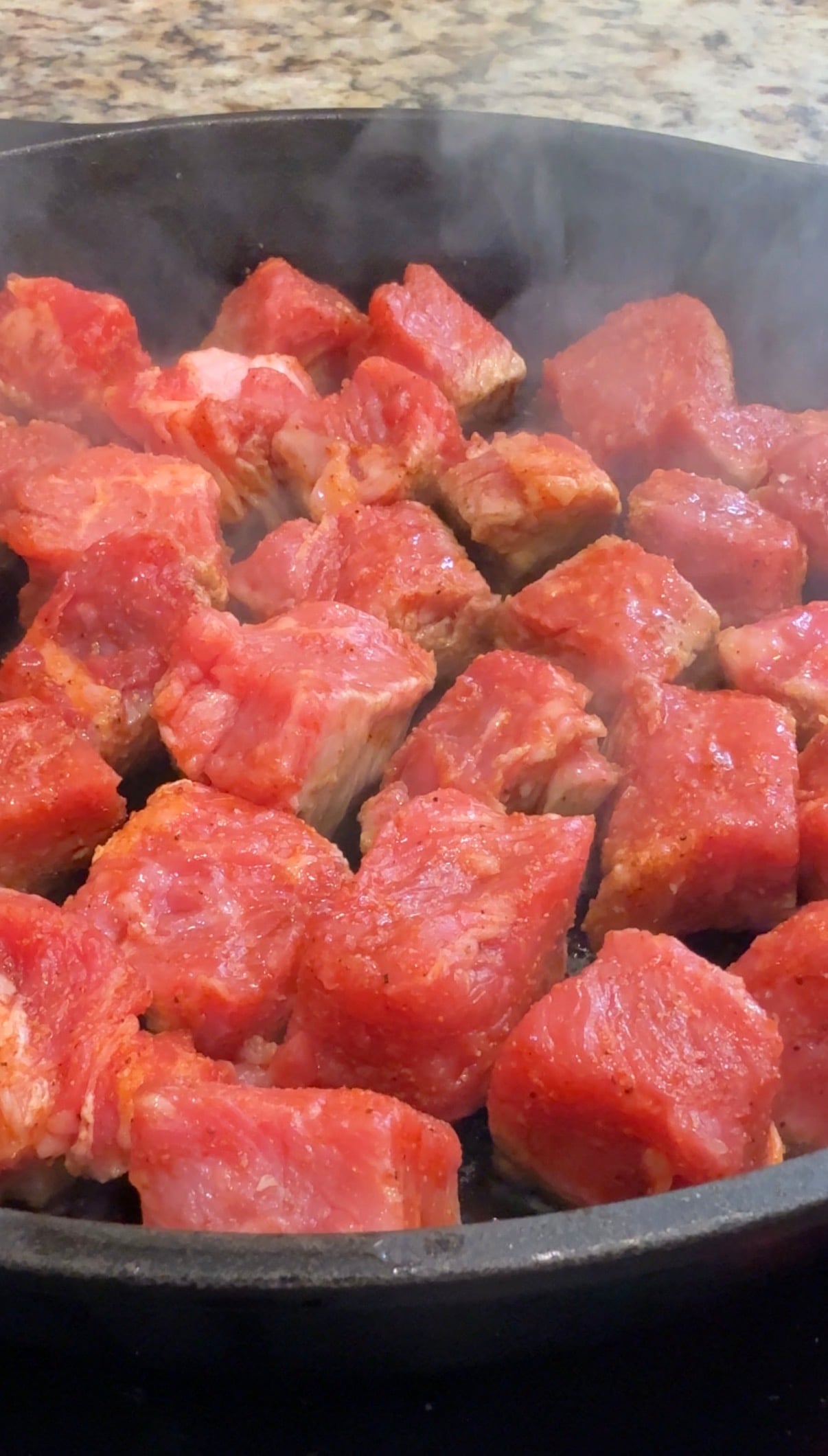 Chunks of raw beef are sizzling and browning in a hot pan, with steam rising, indicating the meat is being seared or cooked.