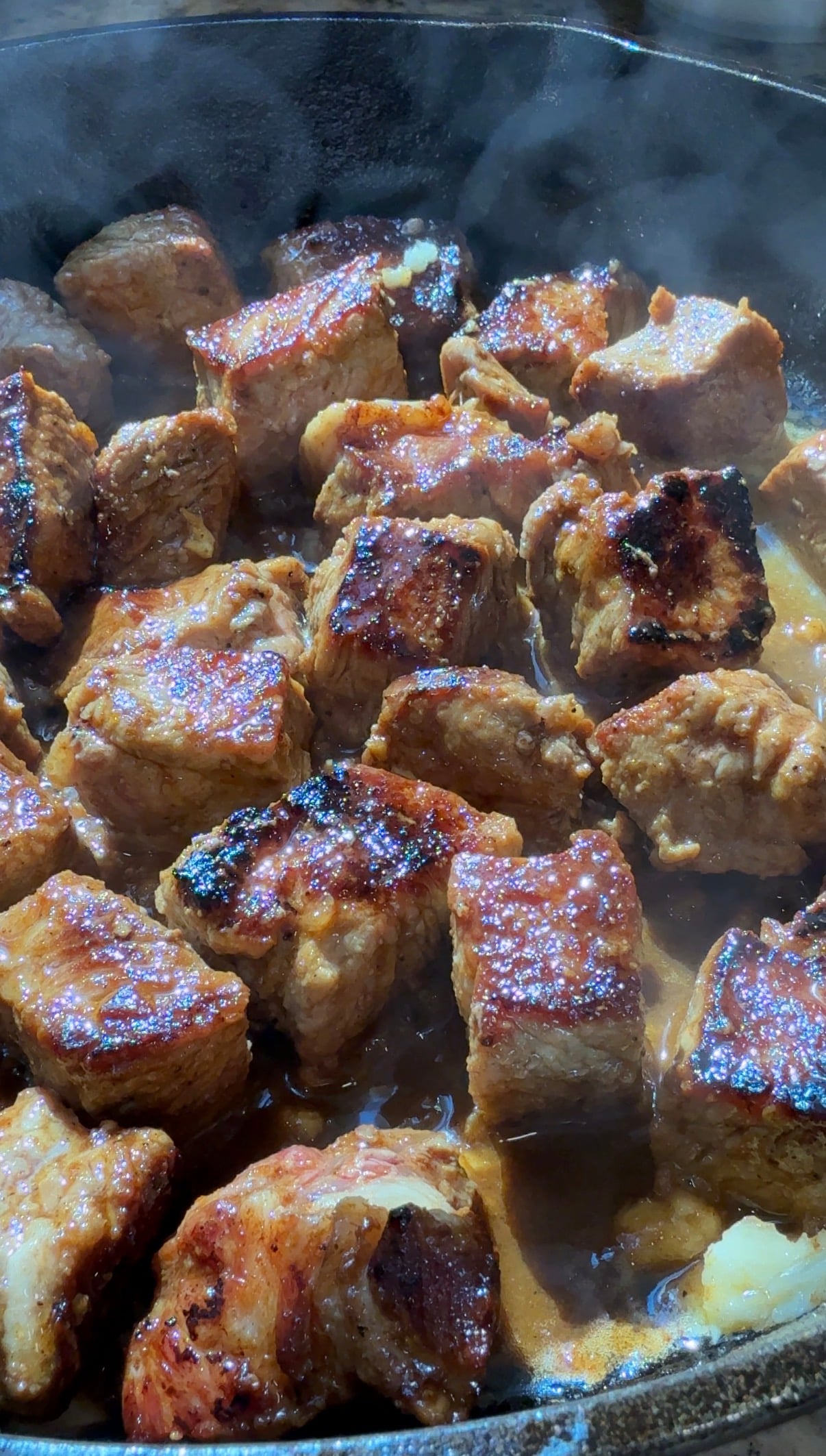 Close-up of browned, seared cubes of meat cooking in a pan, with caramelized edges and a glossy, savory sauce. Steam rises from the hot dish, highlighting the juicy, tender texture of the meat.