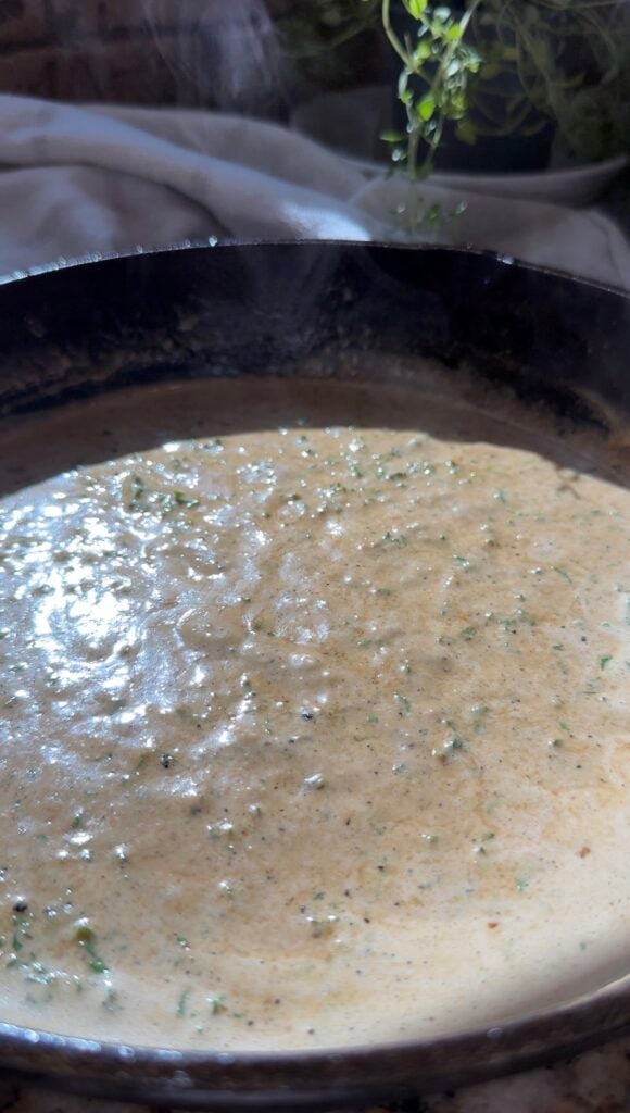 A creamy sauce with herbs simmers in a black cast iron pan, reflecting light on its surface.