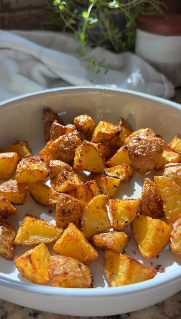 A bowl of golden, crispy roasted potato cubes sits in sunlight, with a white cloth and hints of greenery in the blurred background.