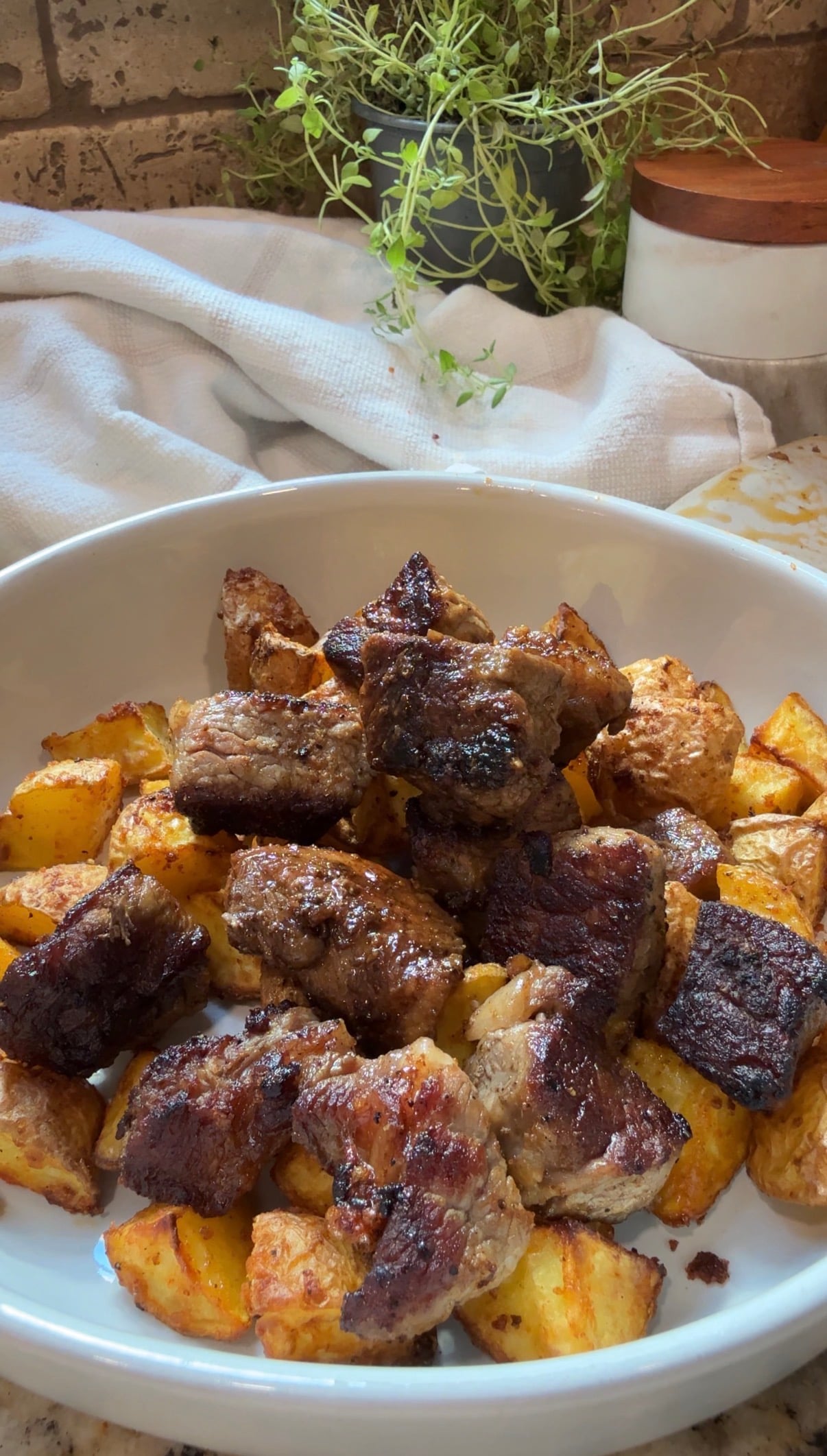 A white bowl filled with golden roasted potatoes and browned, grilled meat cubes sits on a table with a white cloth and fresh herbs in the background.