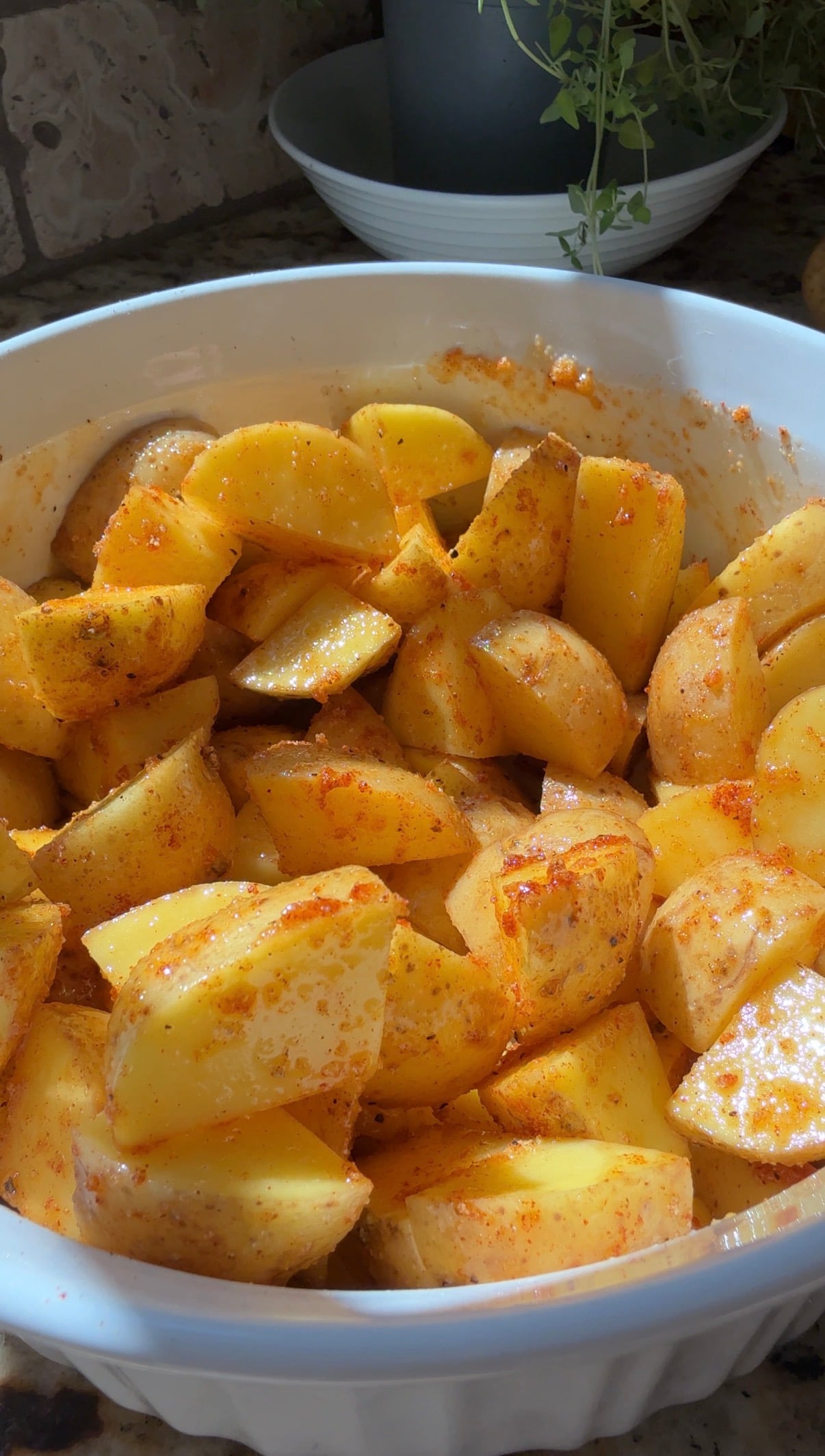 A white bowl filled with chopped potatoes coated in spices, ready to be cooked. Sunlight highlights the yellow and orange hues on the potatoes.