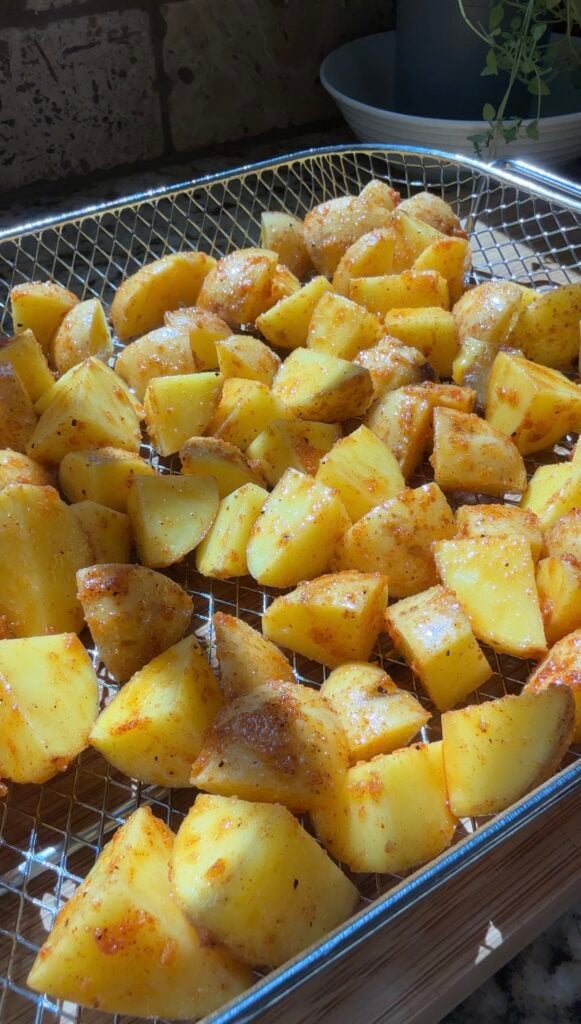 Cubed potatoes with seasoning are spread out on a metal wire rack, ready for roasting or air frying. Sunlight highlights the yellow color and spices on the potatoes.