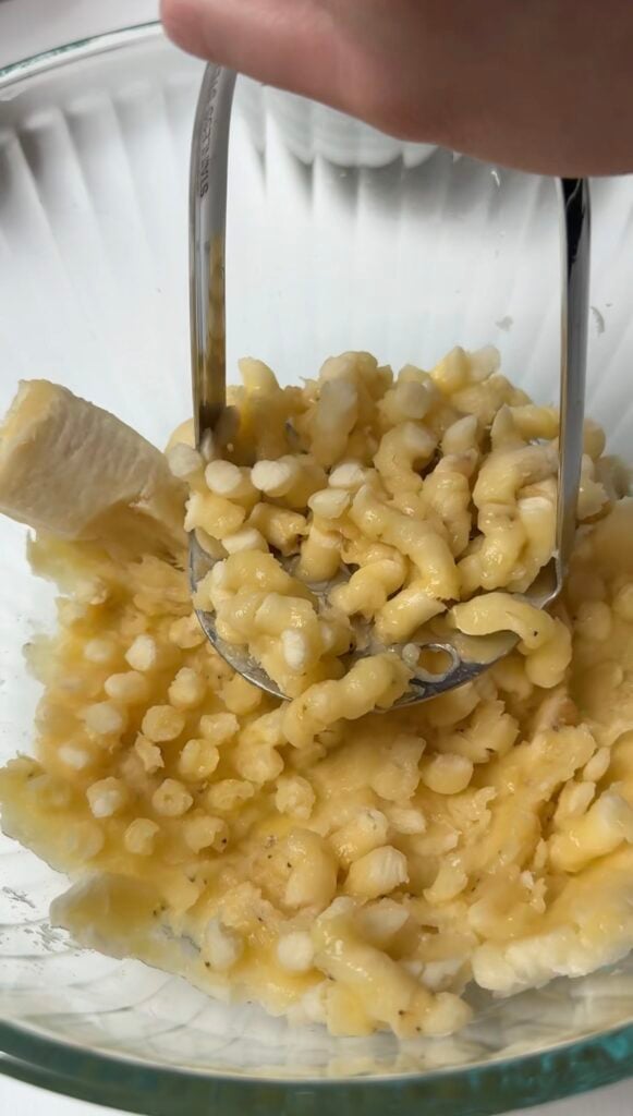 A hand uses a metal masher to mash bananas in a clear glass bowl. The bananas are partially mashed, with chunks and smooth parts visible.