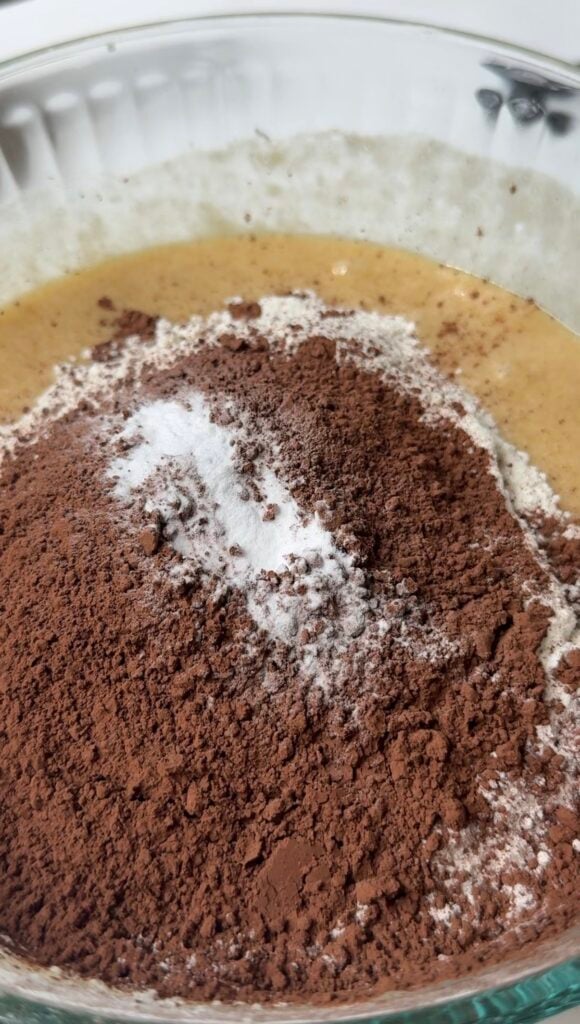 A close-up of a mixing bowl with cocoa powder, flour, baking powder, and sugar on top of a wet yellow batter, ready to be mixed.