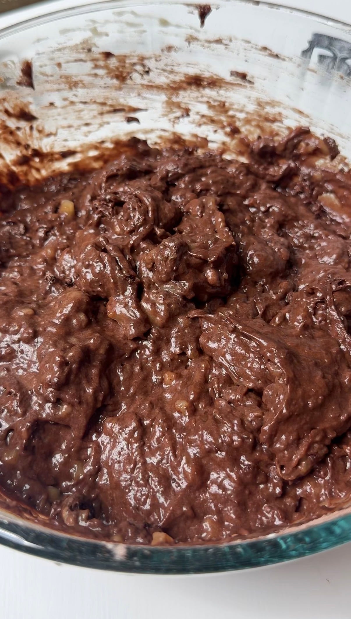 A close-up of thick, chunky chocolate brownie batter in a glass mixing bowl, showing a rich, dark brown color and a slightly glossy texture.