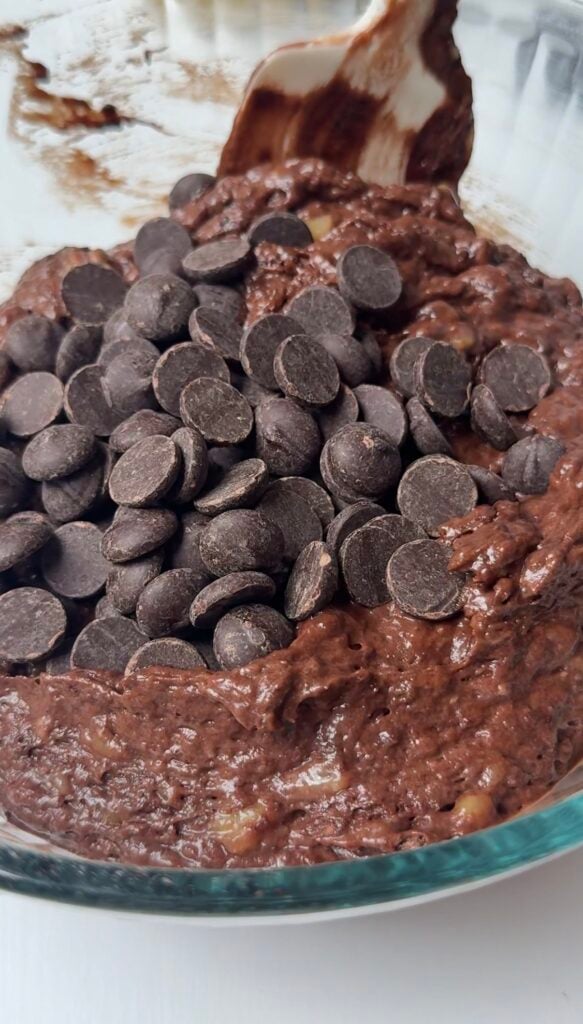 A close-up of chocolate chips on top of thick chocolate batter in a glass mixing bowl, ready to be mixed together.