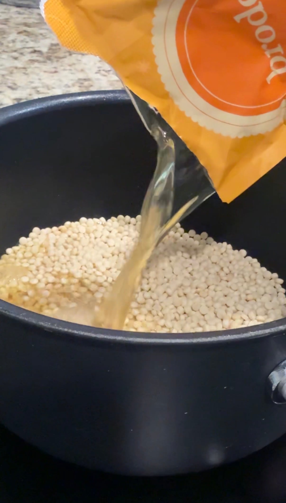 Broth is being poured from a carton into a pot filled with uncooked white orzo pasta.