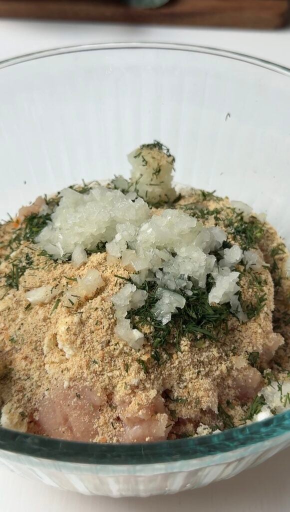 A close-up of a bowl containing ground meat mixed with breadcrumbs, chopped onions, and herbs, ready for preparation.