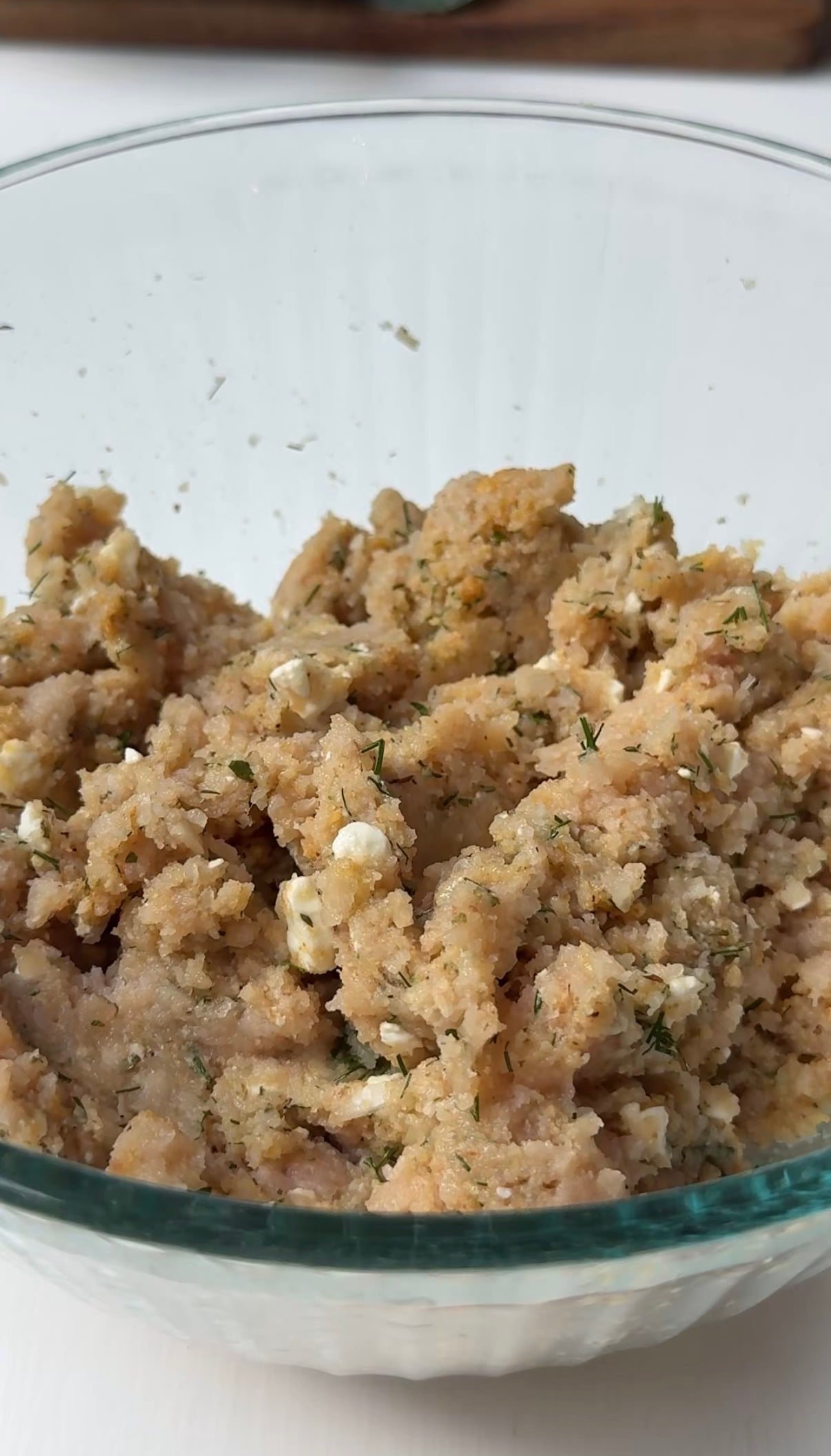 A close-up of a glass bowl filled with a mixture of ground chicken, herbs, breadcrumbs, and crumbled cheese, ready for cooking.