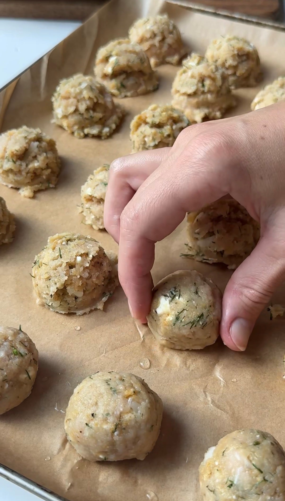 A hand forms a ball of uncooked stuffing mixture on a baking sheet lined with parchment paper. Several other stuffing balls are arranged in the background, ready to be baked.