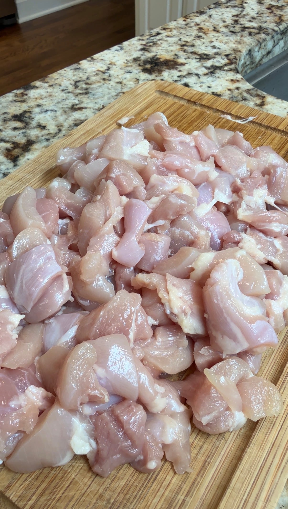 A pile of raw, diced chicken pieces on a wooden cutting board, with a granite countertop in the background.