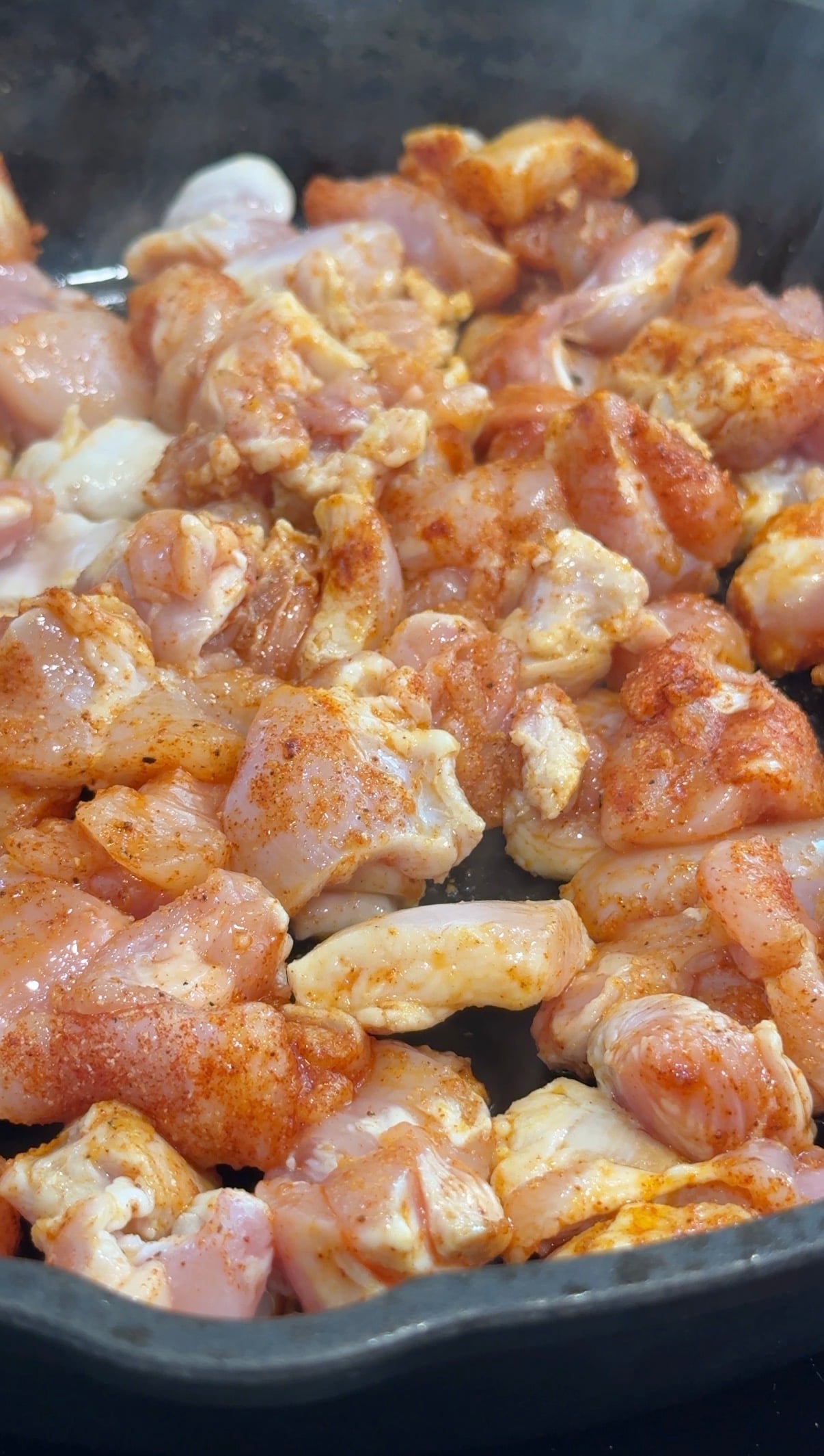 Close-up of raw, seasoned chicken pieces cooking in a pan, with visible spices and some steam rising.