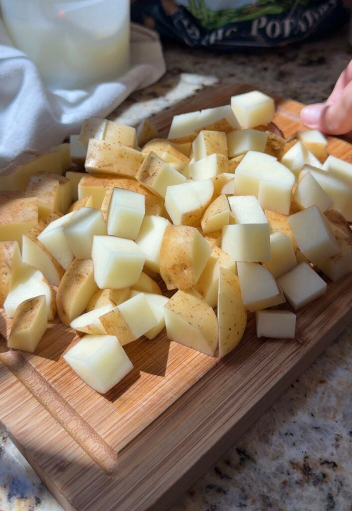 Diced potatoes with skins on are spread out on a wooden cutting board. A hand is visible in the corner, and the scene is lit by natural sunlight.