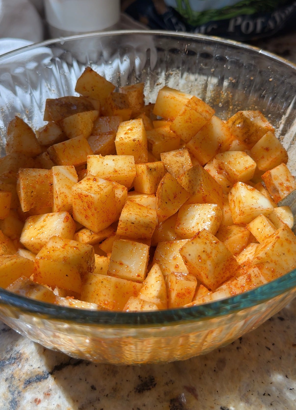 A glass bowl filled with cubed potatoes seasoned with red spices, likely paprika, sits on a granite countertop in bright sunlight.