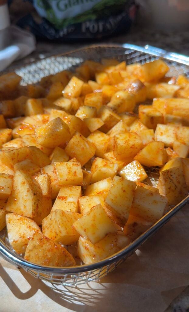 Diced potatoes coated with spices are arranged in a metal air fryer basket, ready for cooking. Sunlight highlights the golden color and seasoning on the potato pieces.