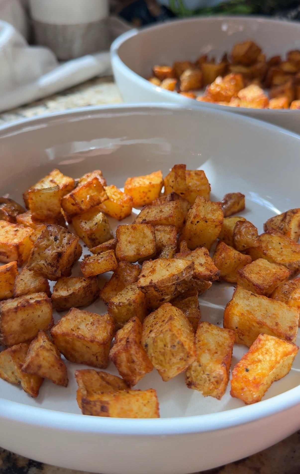 Golden, crispy roasted potato cubes seasoned with spices are served in a white dish, with another similar dish in the background on a kitchen counter, sunlight highlighting their texture.