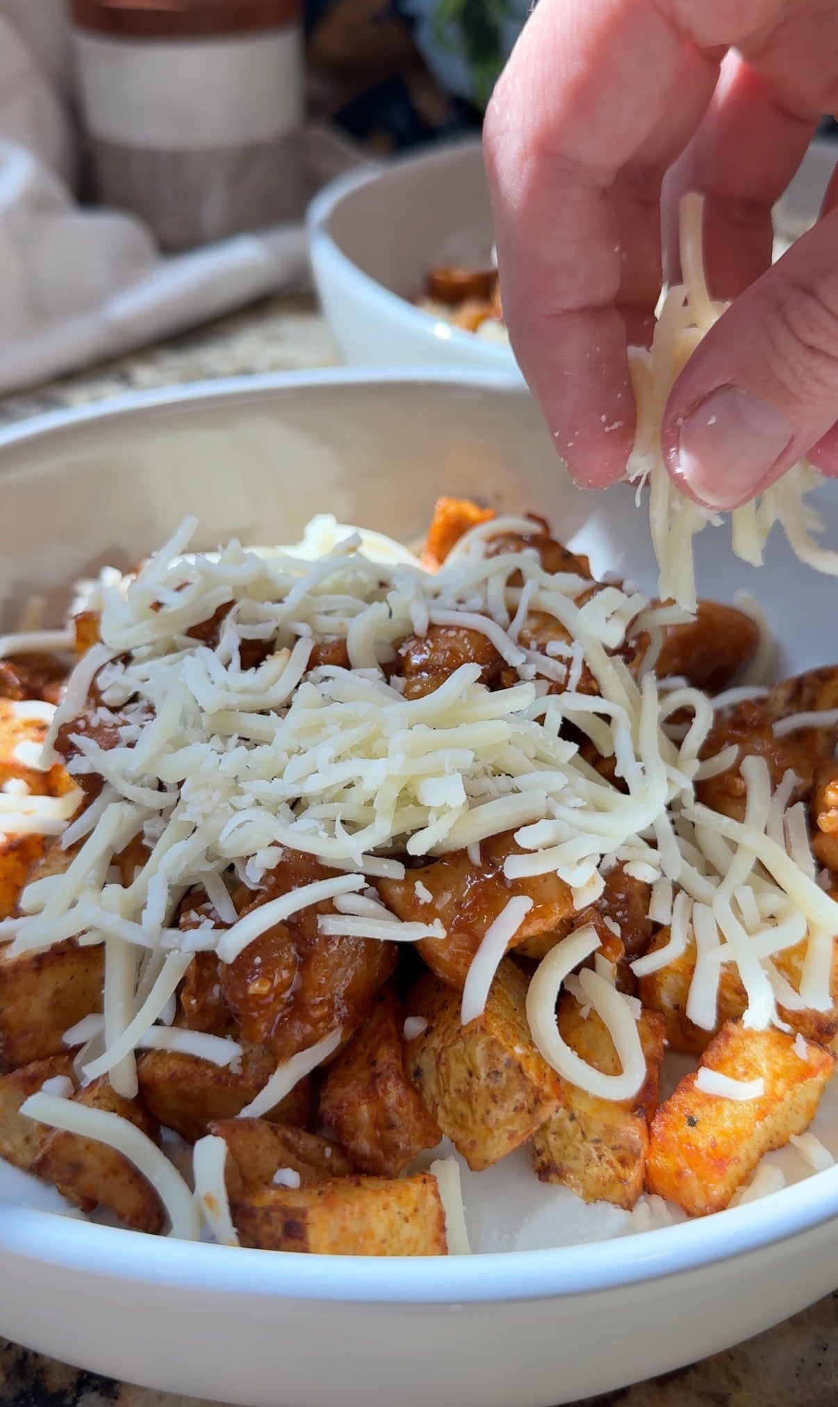 A hand sprinkles shredded cheese over a bowl of seasoned potato wedges topped with chili, with another bowl visible in the background.