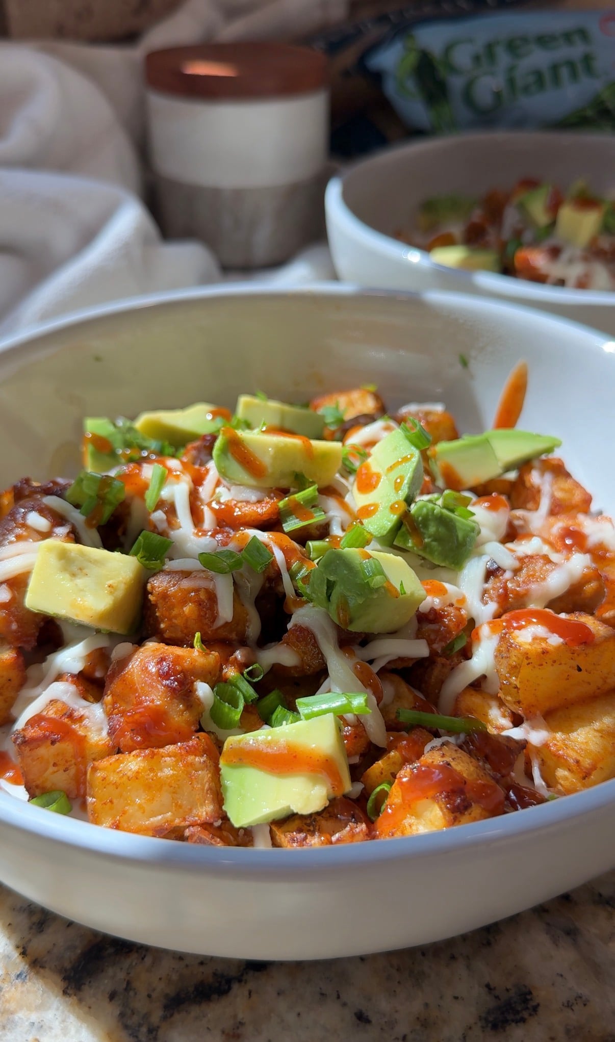 A white bowl filled with diced potatoes, topped with avocado cubes, chopped green onions, drizzled with sauce and white dressing, on a granite countertop with another bowl and seasoning in the background.
