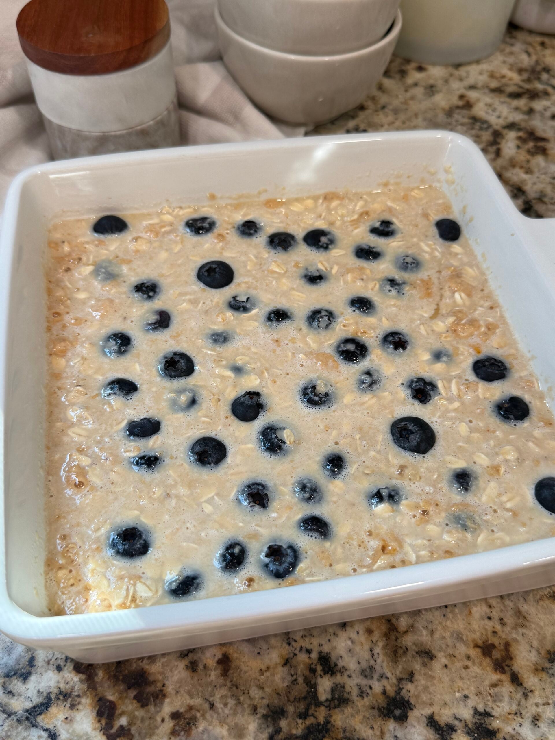 A white baking dish filled with unbaked oatmeal mixture, topped with scattered fresh blueberries, sits on a granite countertop. Bowls and jars are in the background.