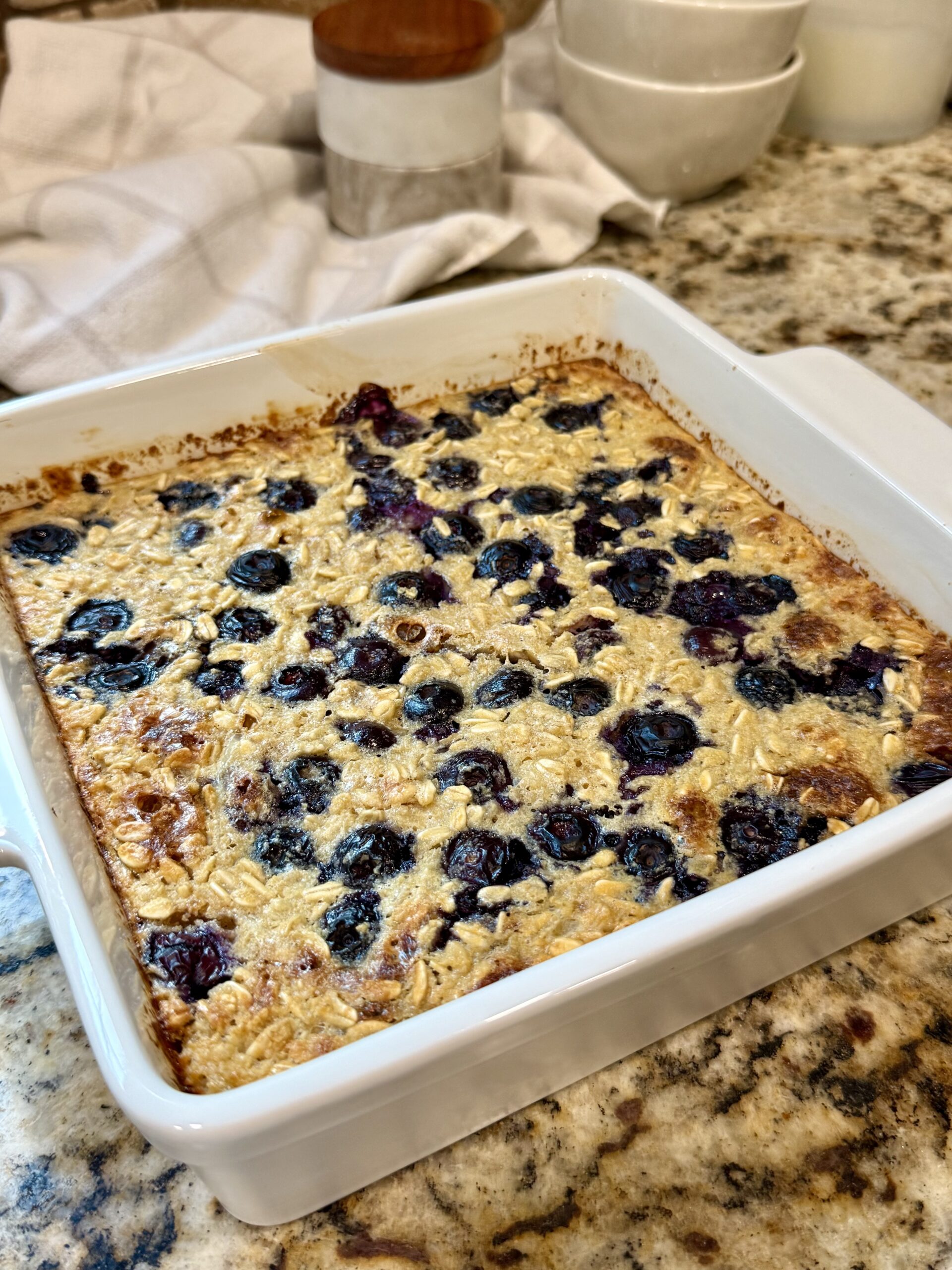 A baked oatmeal with blueberries in a white square dish sits on a kitchen counter, with bowls and containers in the background.
