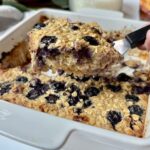 A hand holds a spatula lifting a square of baked oatmeal with blueberries from a white baking dish. The oatmeal looks moist and studded with berries.