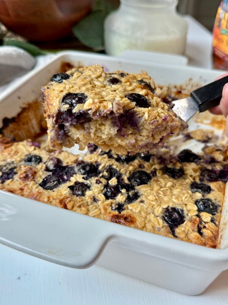 A hand holds a spatula lifting a square of baked oatmeal with blueberries from a white baking dish. The oatmeal looks moist and studded with berries.