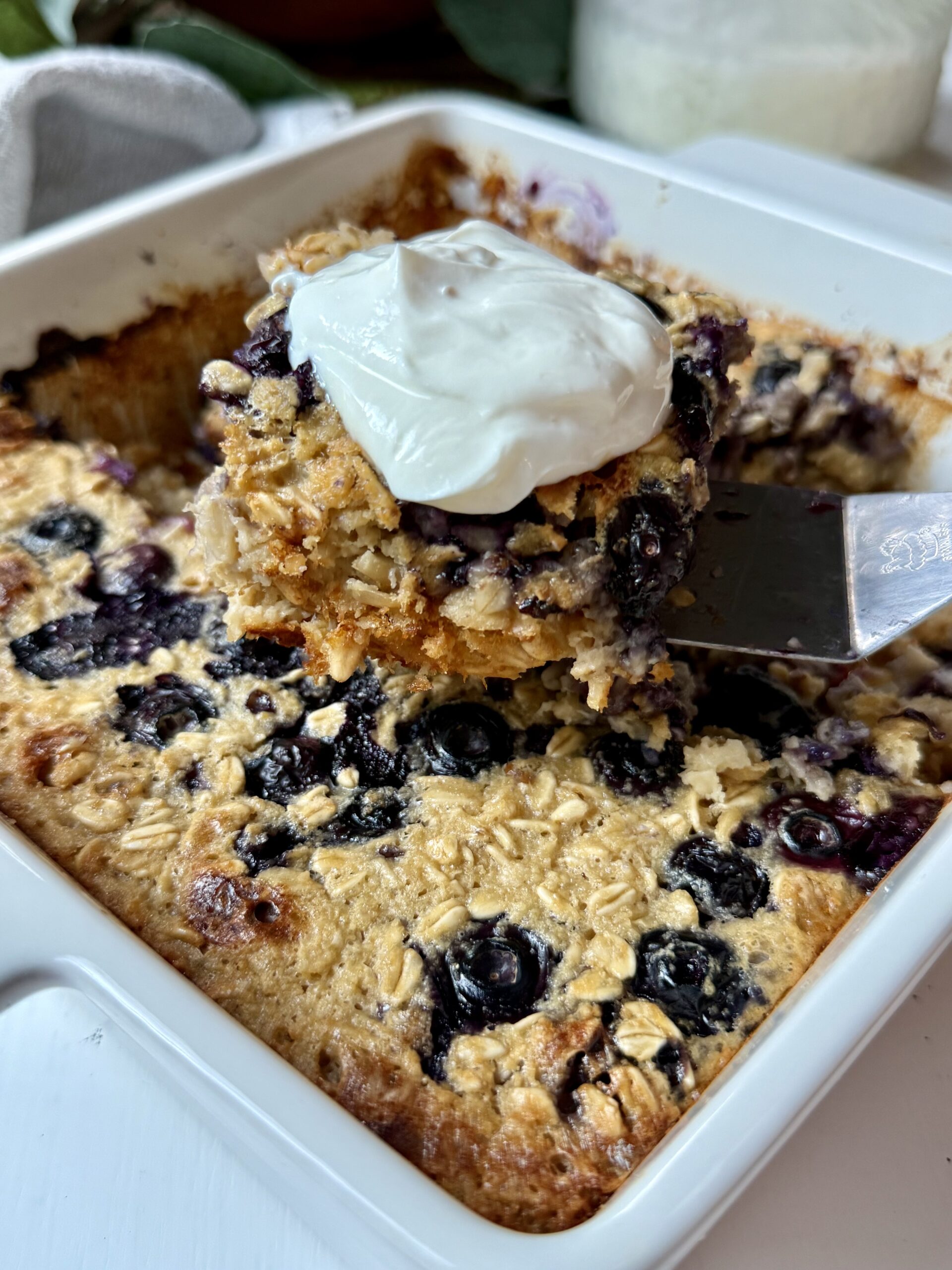 A spatula lifts a serving of baked oatmeal topped with yogurt from a white square dish filled with blueberry oatmeal bake.