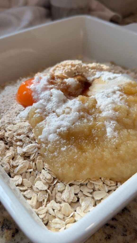 A close-up of a bowl with oats, mashed banana, an egg yolk, baking powder, and vanilla extract, ready to be mixed for baking.