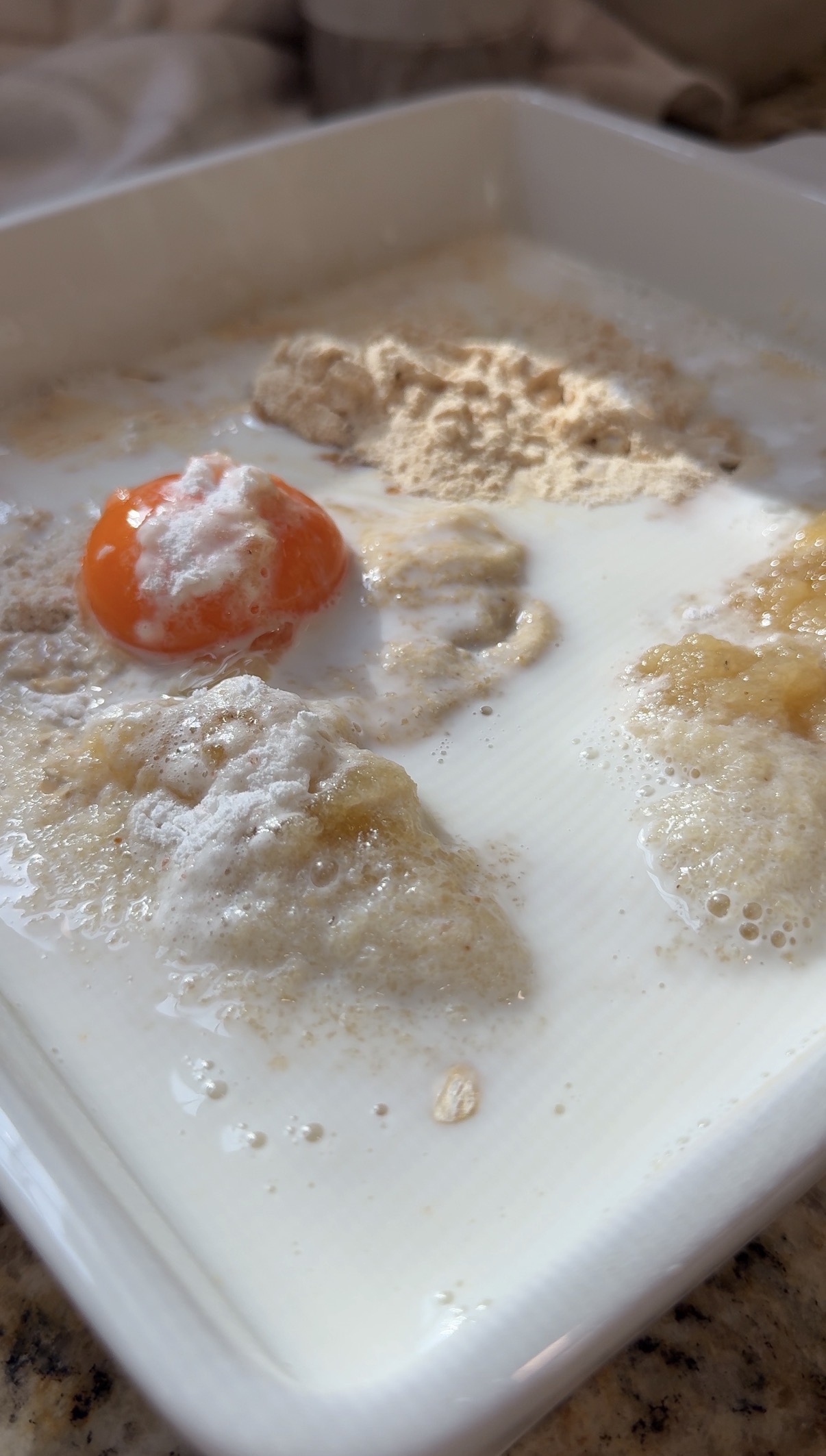 Close-up of raw baking ingredients in a bowl, including an egg yolk, flour, oats, banana, and milk, before mixing.