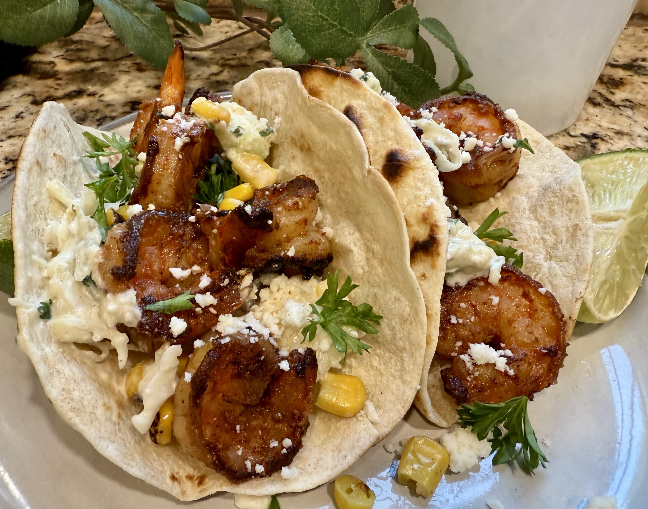 Two shrimp tacos with grilled shrimp, corn, cabbage slaw, crumbled cheese, and fresh herbs on flour tortillas, served with lime wedges on a plate.