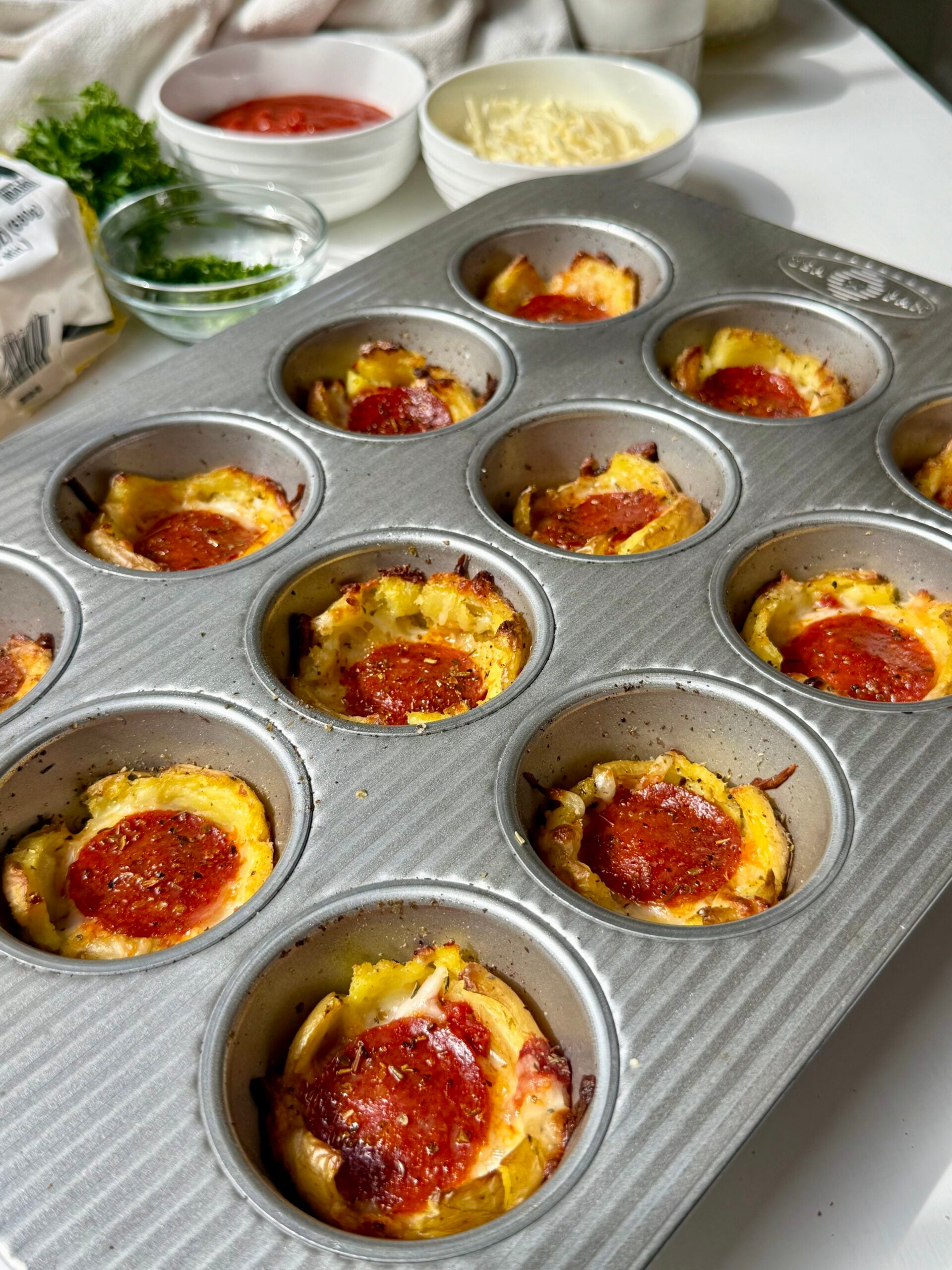 Muffin tin filled with mini pepperoni pizzas, topped with melted cheese and pepperoni slices. Bowls of sauce, shredded cheese, and parsley are in the background on a white surface.