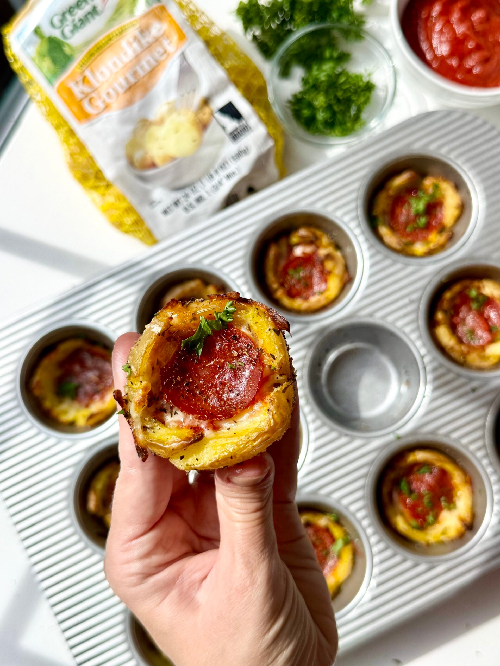 A hand holds a muffin tin pizza bite topped with pepperoni and parsley. Other pizza bites are in a muffin pan. Shredded potatoes, parsley, and pizza sauce are nearby.