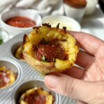 A hand holds a baked potato cup filled with cheese and a slice of pepperoni above a muffin tin, with bowls of sauce and toppings in the background.