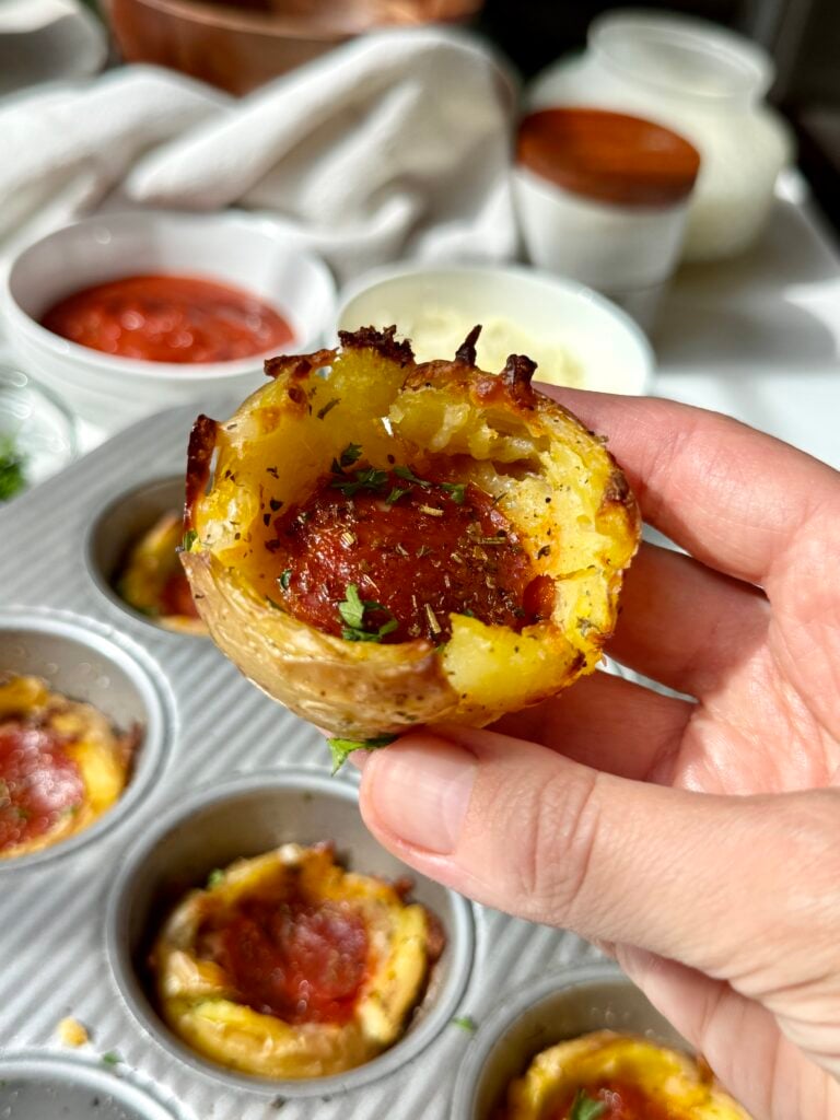 A hand holds a baked potato cup filled with cheese and a slice of pepperoni above a muffin tin, with bowls of sauce and toppings in the background.