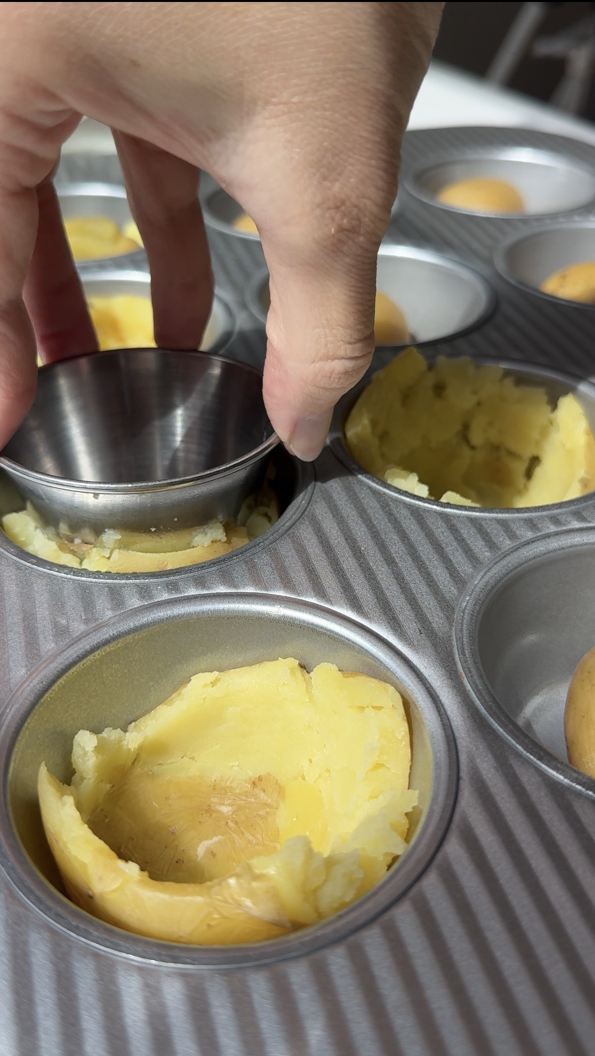 A hand presses a metal cup into a muffin tin filled with dough, shaping it into small crusts. Unshaped dough is visible in some cups, while others are already molded.