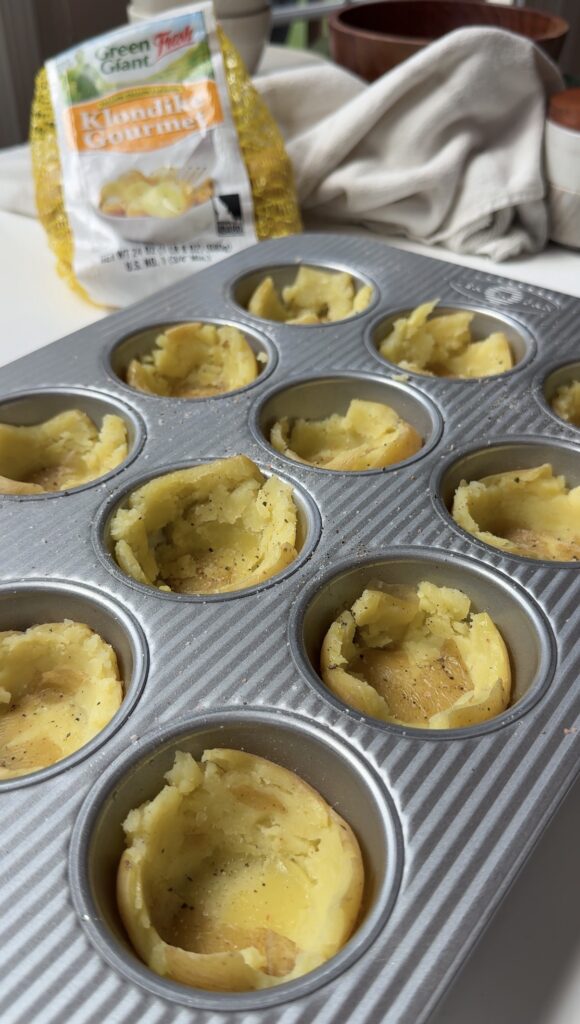 A muffin tin filled with partially hollowed baked potato cups sits on a counter. In the background, there is a bag of Klondike Gourmet potatoes and a white kitchen towel.