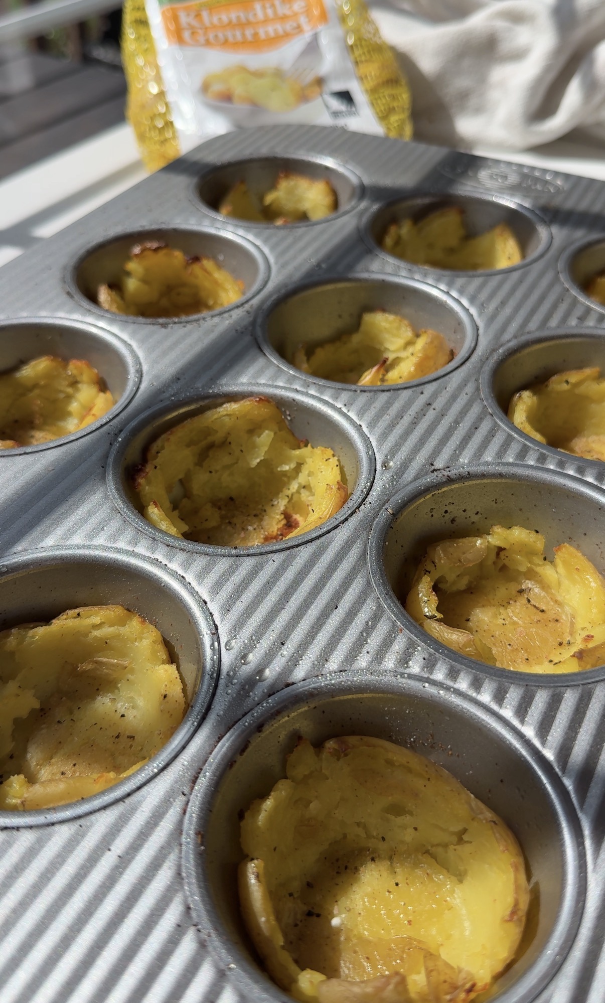 A muffin tin filled with partially hollowed baked potato cups sits on a counter. In the background, there is a bag of Klondike Gourmet potatoes and a white kitchen towel.