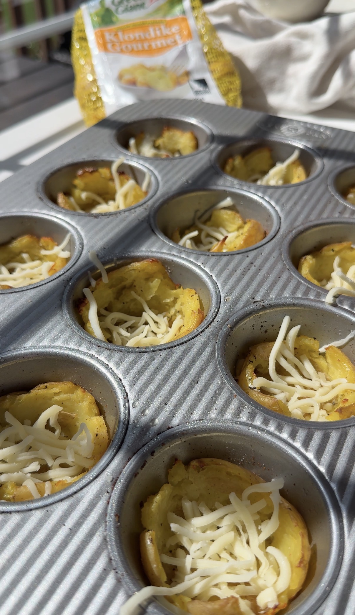 Muffin tin with baked yellow potato cups topped with shredded cheese, sitting on a sunny countertop. A bag labeled Klondike Gourmet with small yellow potatoes is visible in the background.