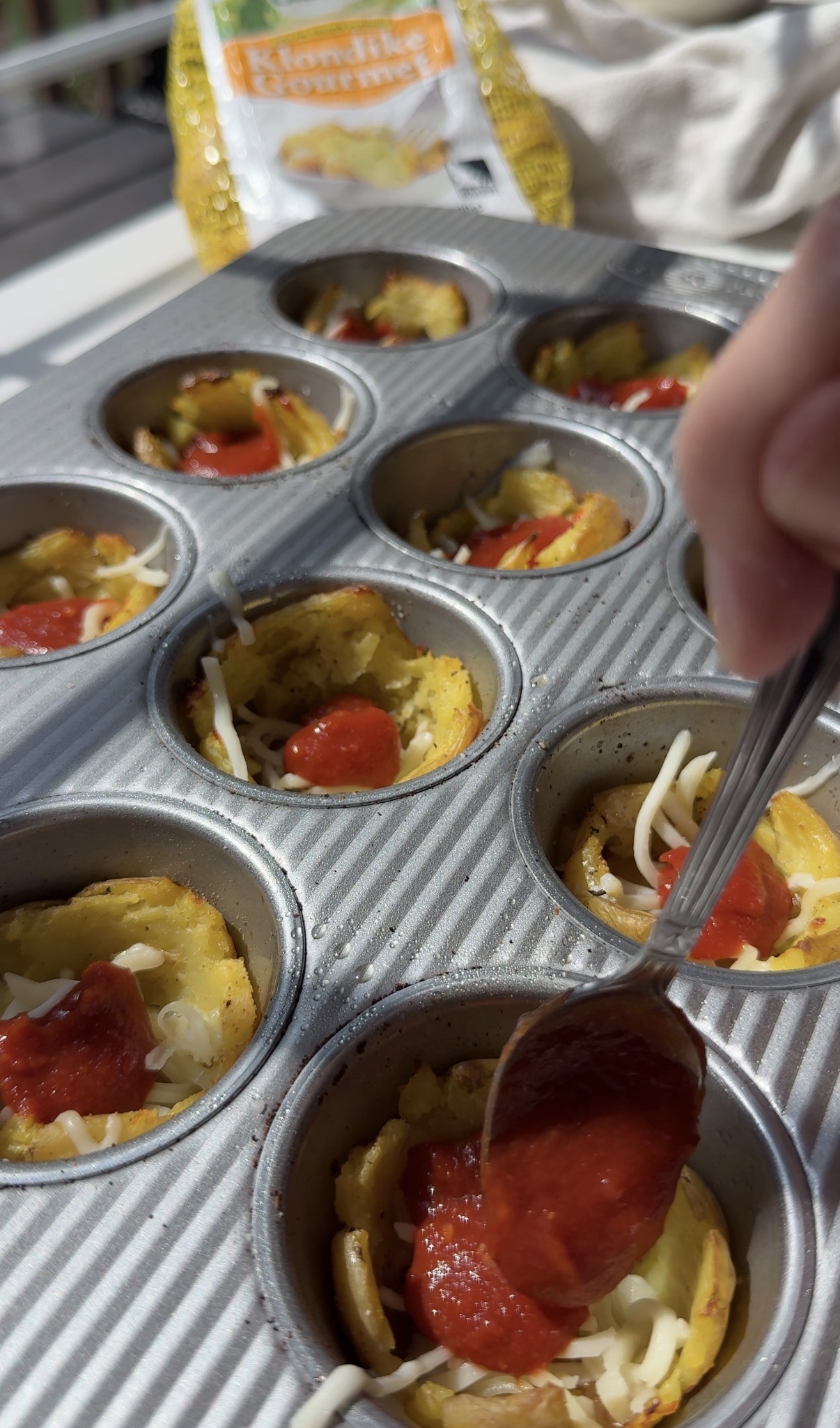 A hand adds pepperoni to mini pizza cups in a metal muffin pan. Shredded cheese and tomato sauce are visible on top of the cups.