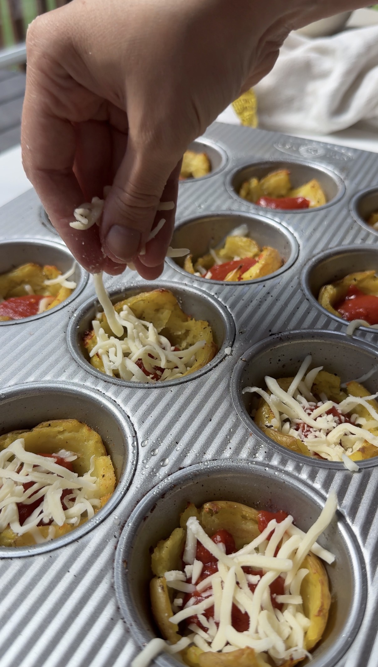 A hand sprinkles shredded cheese over mini omelets with vegetables in a metal muffin tray before baking.