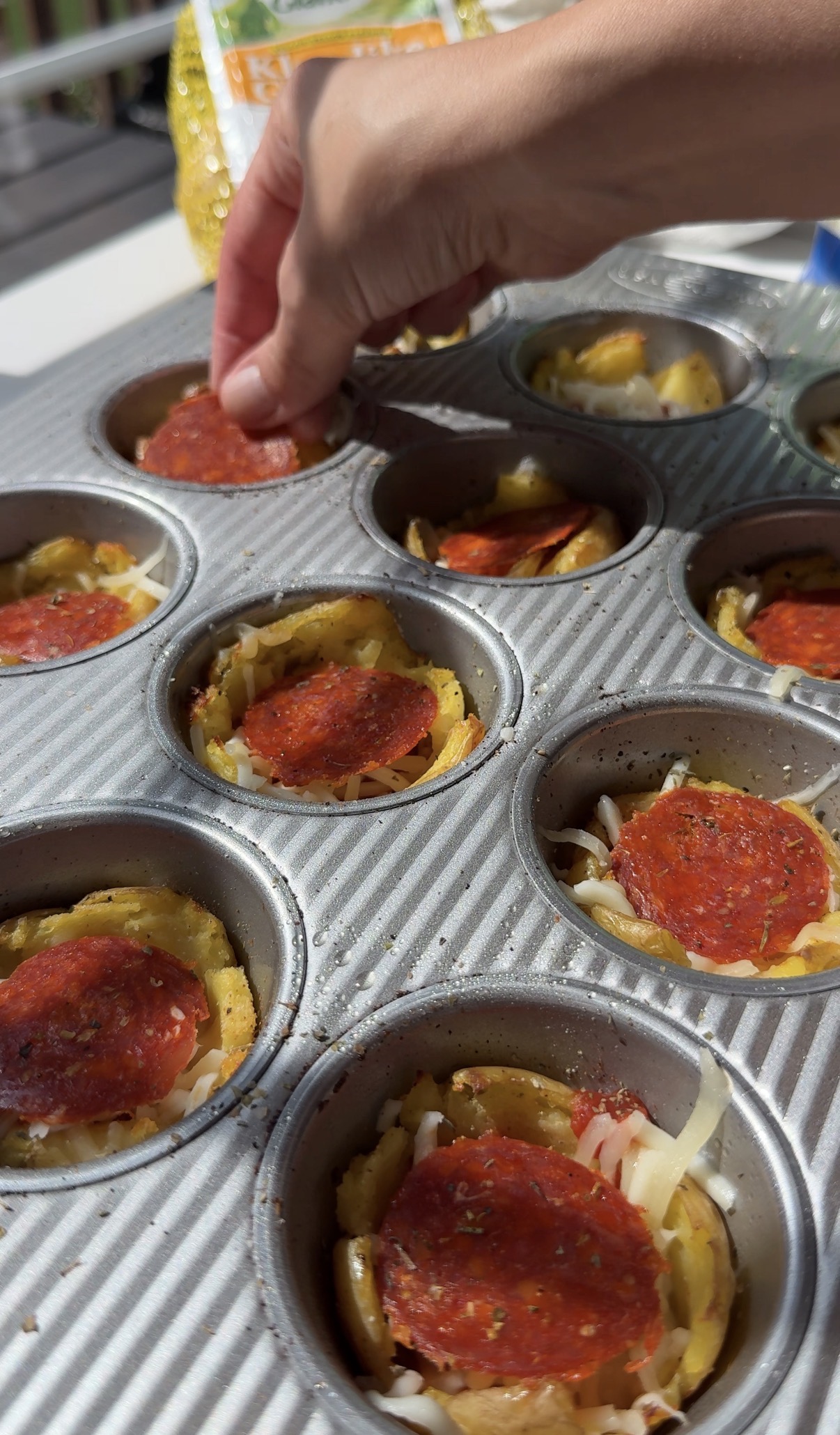 A hand places pepperoni slices on mini pizza cups in a muffin tray, each filled with cheese and other toppings, outdoors in natural light.