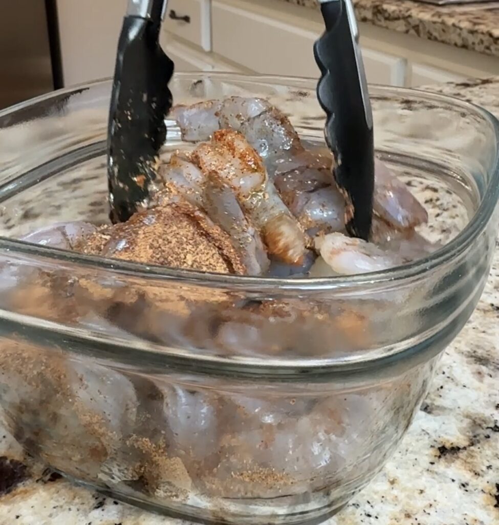 A pair of black tongs mixing raw shrimp with seasoning in a clear glass bowl on a granite kitchen countertop.