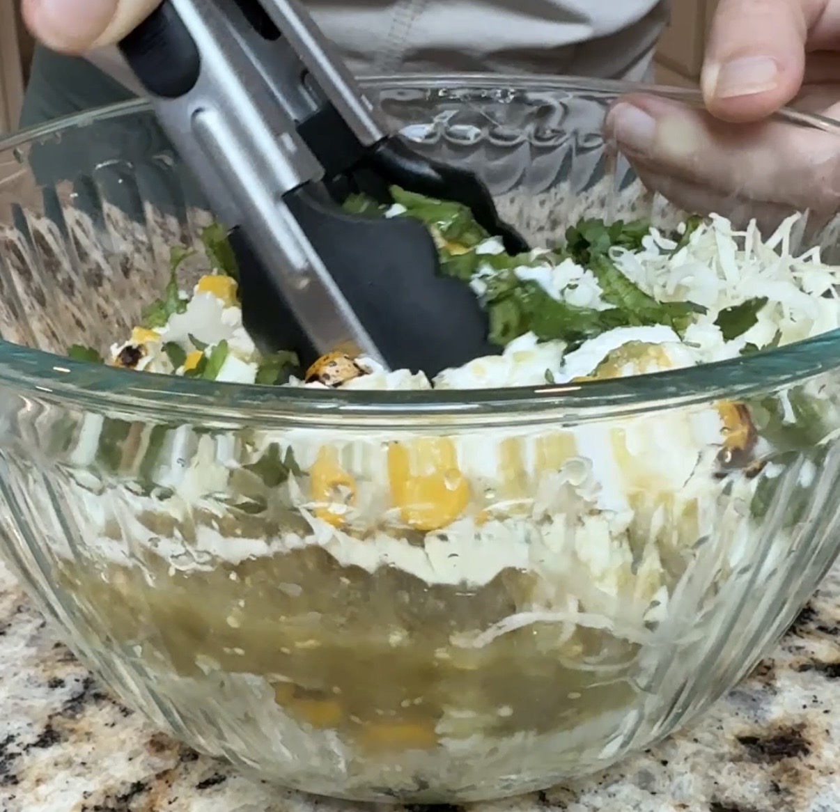 A person uses tongs to mix cheese, cilantro, green salsa, and grilled corn in a glass bowl on a granite countertop.