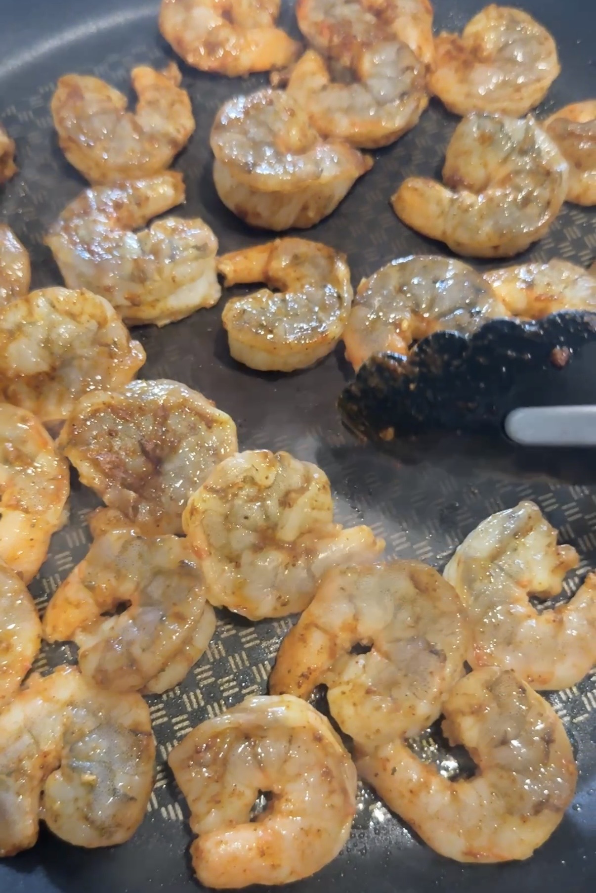 Shrimp seasoned with spices cooking in a frying pan, with a pair of black tongs turning some of the shrimp. The shrimp are orange and appear to be partly cooked.